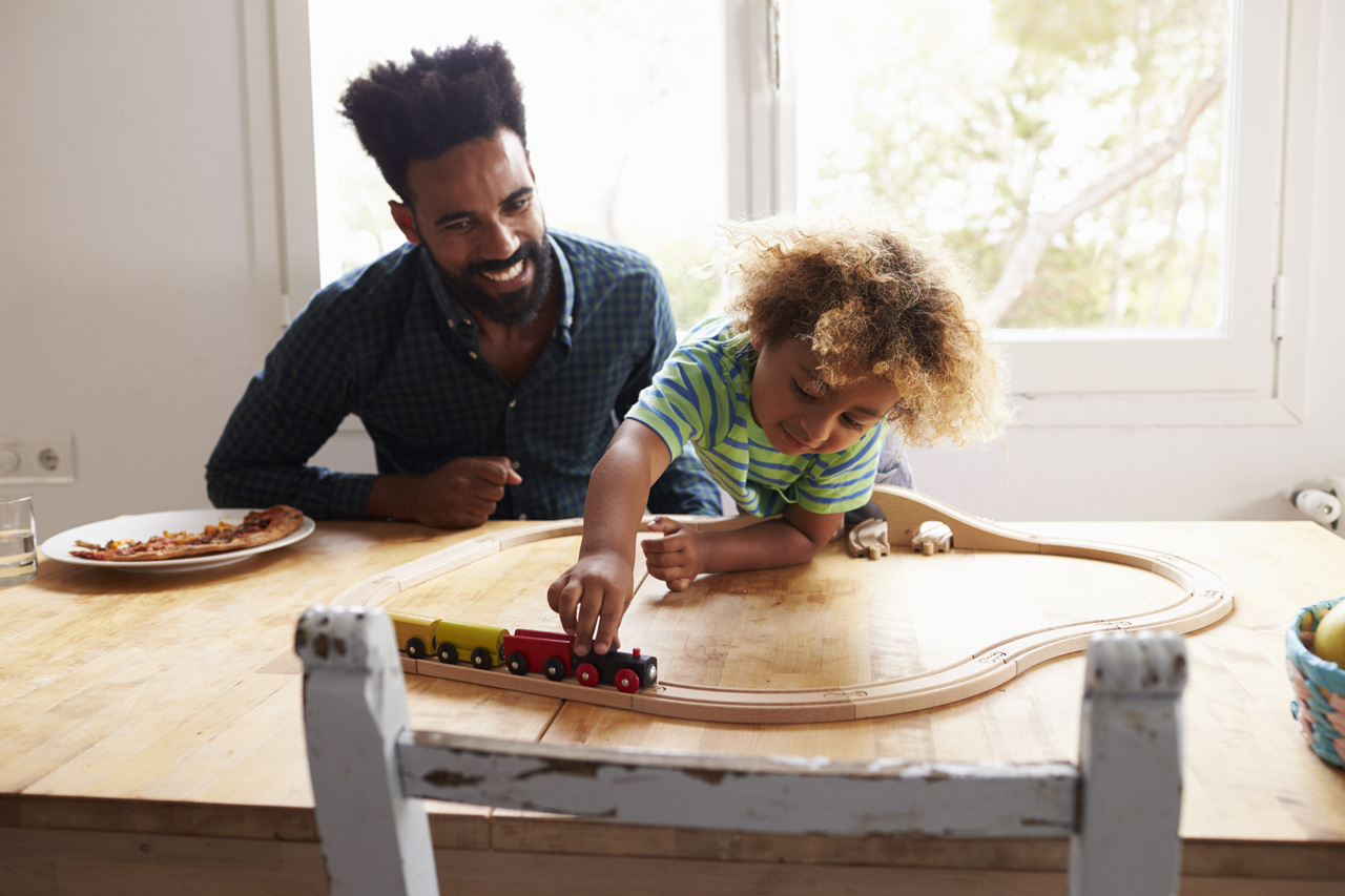 Father and son happily sit at the kitchen table playing with a train set