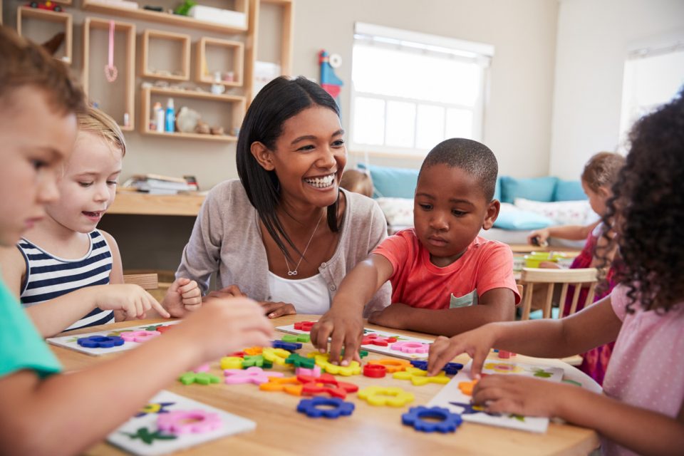 Smiling teacher kneeing at wooden table with children while they work on activities