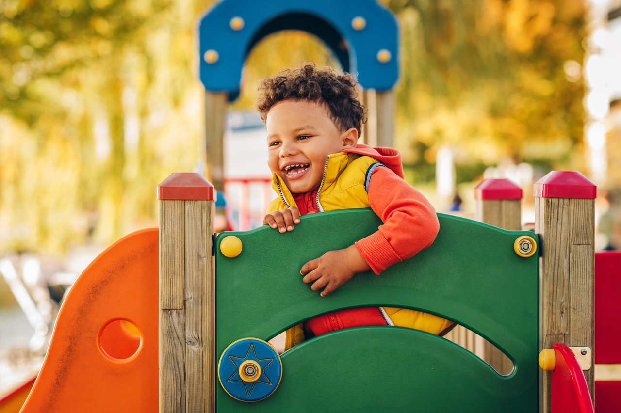 Young boy wearing a yellow vest smilies while playing on green and orange playground