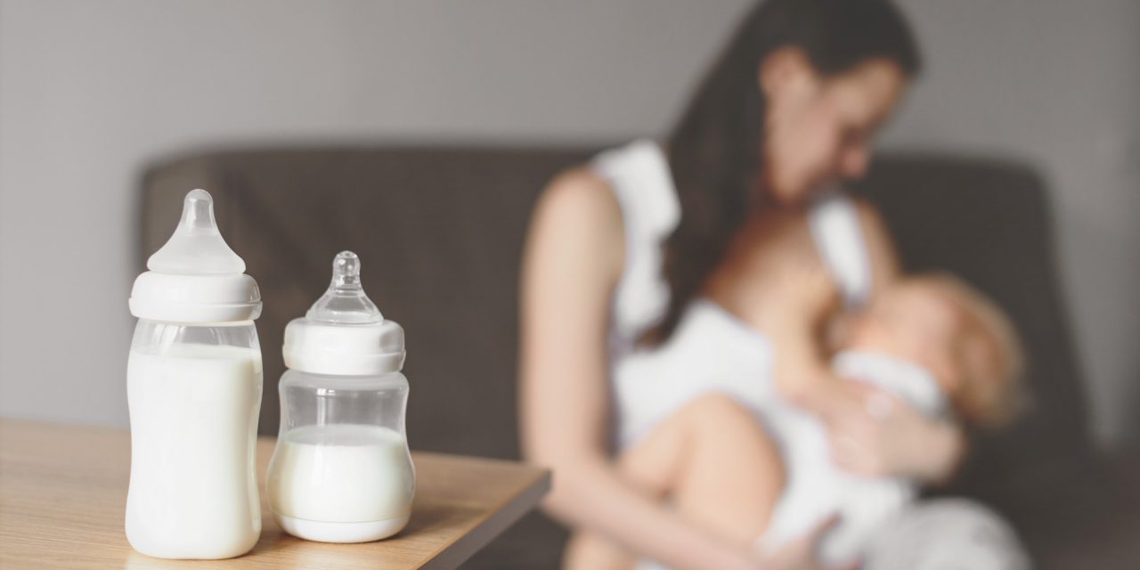 Milk bottles sit on top of wooden table with mom sitting on brown couch breastfeeding baby in the background