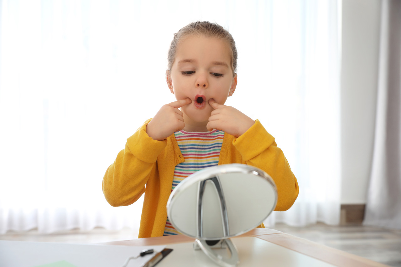 Young girl wearing a yellow sweater looks into the mirror with fingers pointing into cheeks while practicing speech therapy