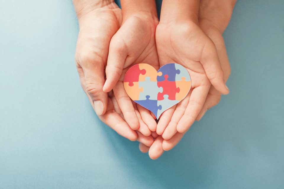 Women and child with hands laying inside of one another hold a colorful heart made out of puzzle pieces