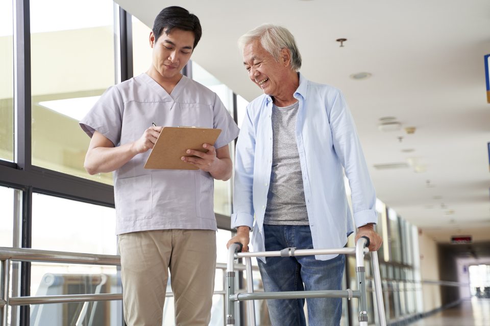 young asian physical therapist working with old man on working using a walker in hallway of nursing home