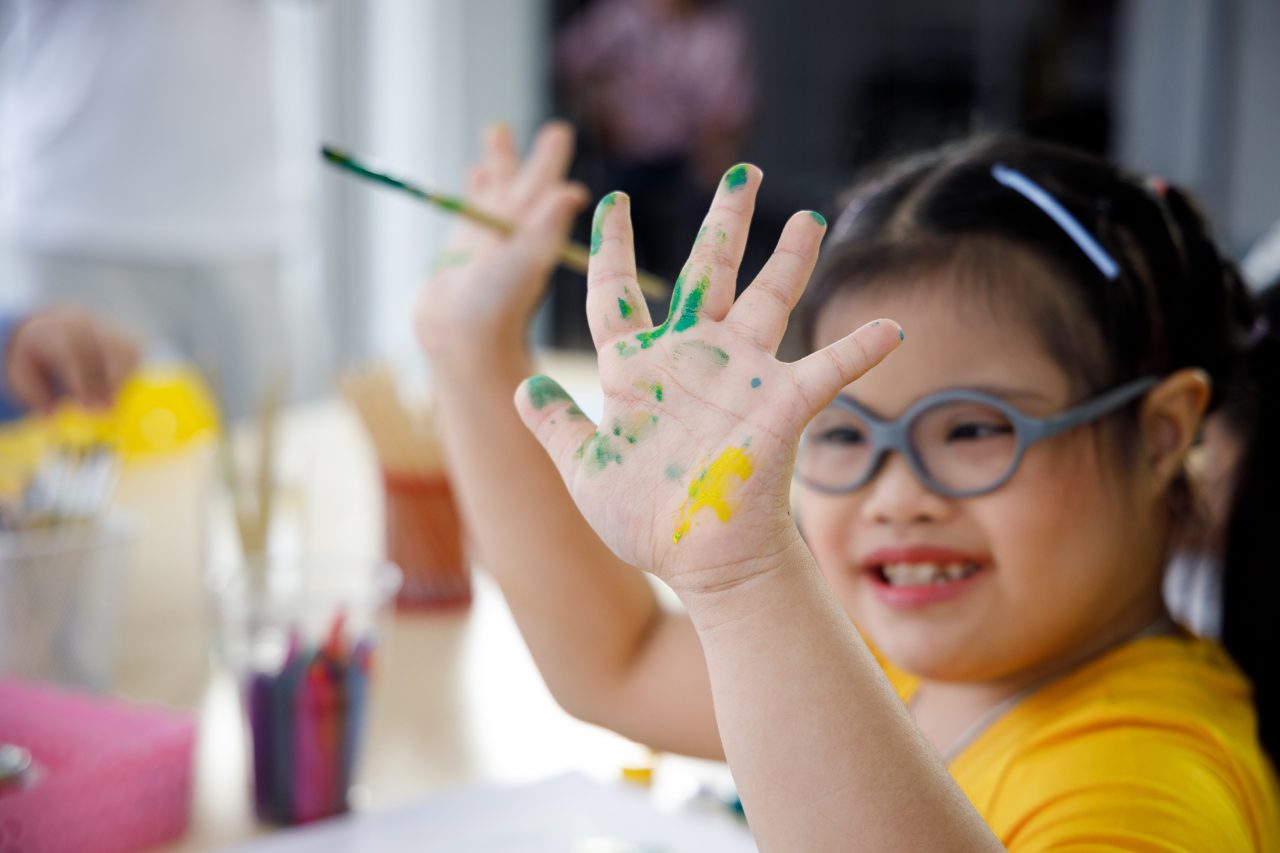 Asian girl in yellow shirt with Down's syndrome painting her hand.