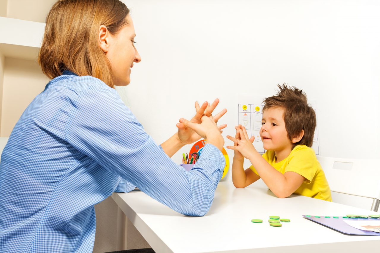 Boy exercises by putting hands and fingers together with therapist showing it improving motor skills sitting opposite at the table indoors