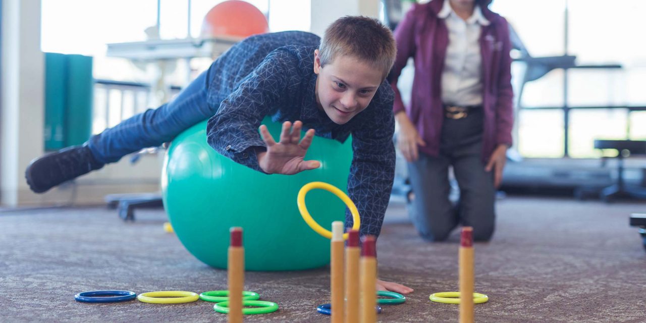 adolescent boy in blue button up shirt laying across large teal exercise ball tossing plastic yellow ring onto ring toss for occupational therapy