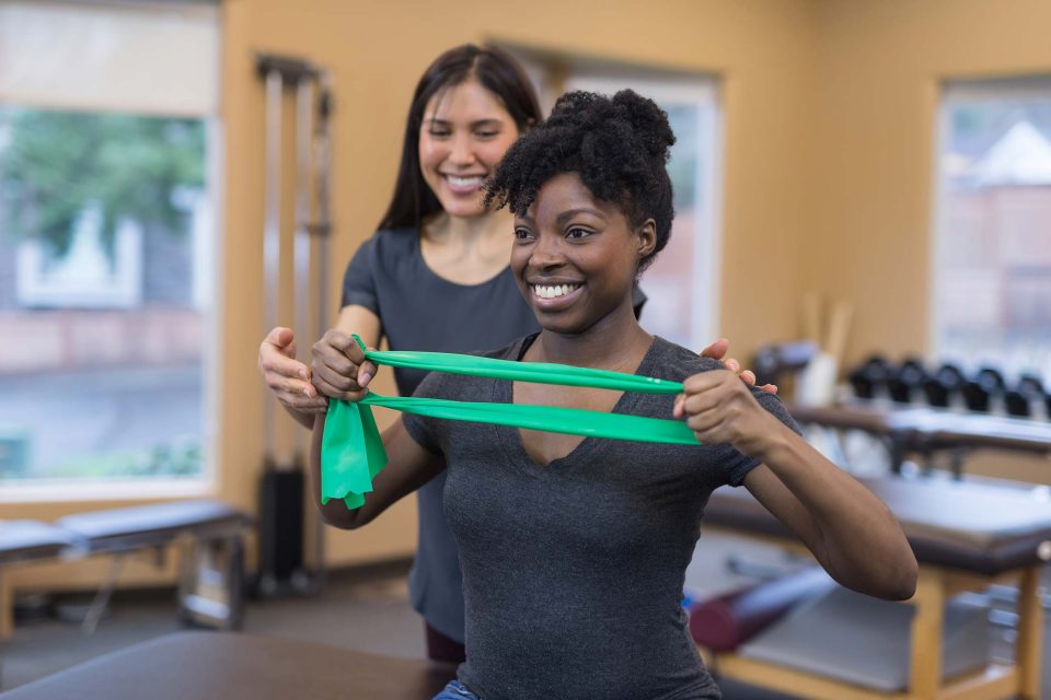 Smiling physical therapist of Asian descent works with her patient, a young African American woman. They are doing a stretching exercise with a flexible green resistance band