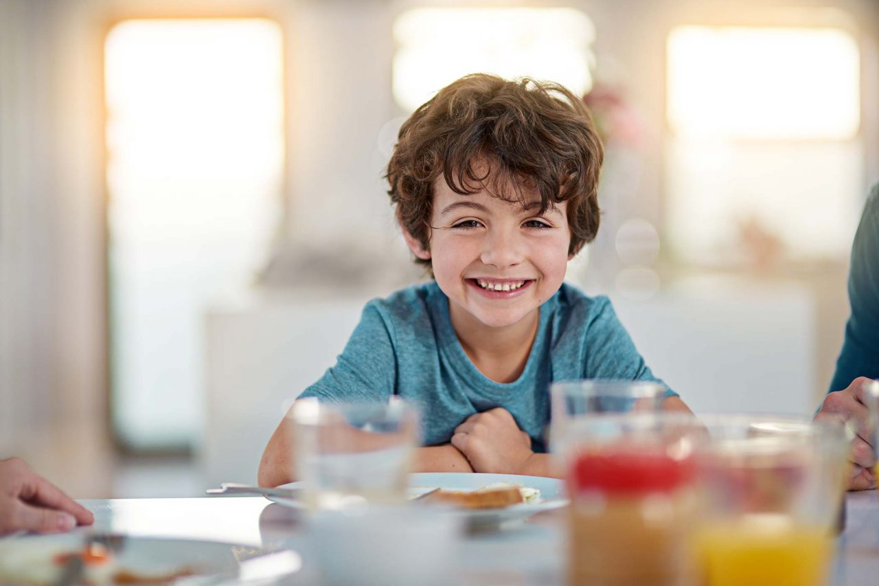 young boy in teal crew neck with tousled brown hair smiles over breakfast