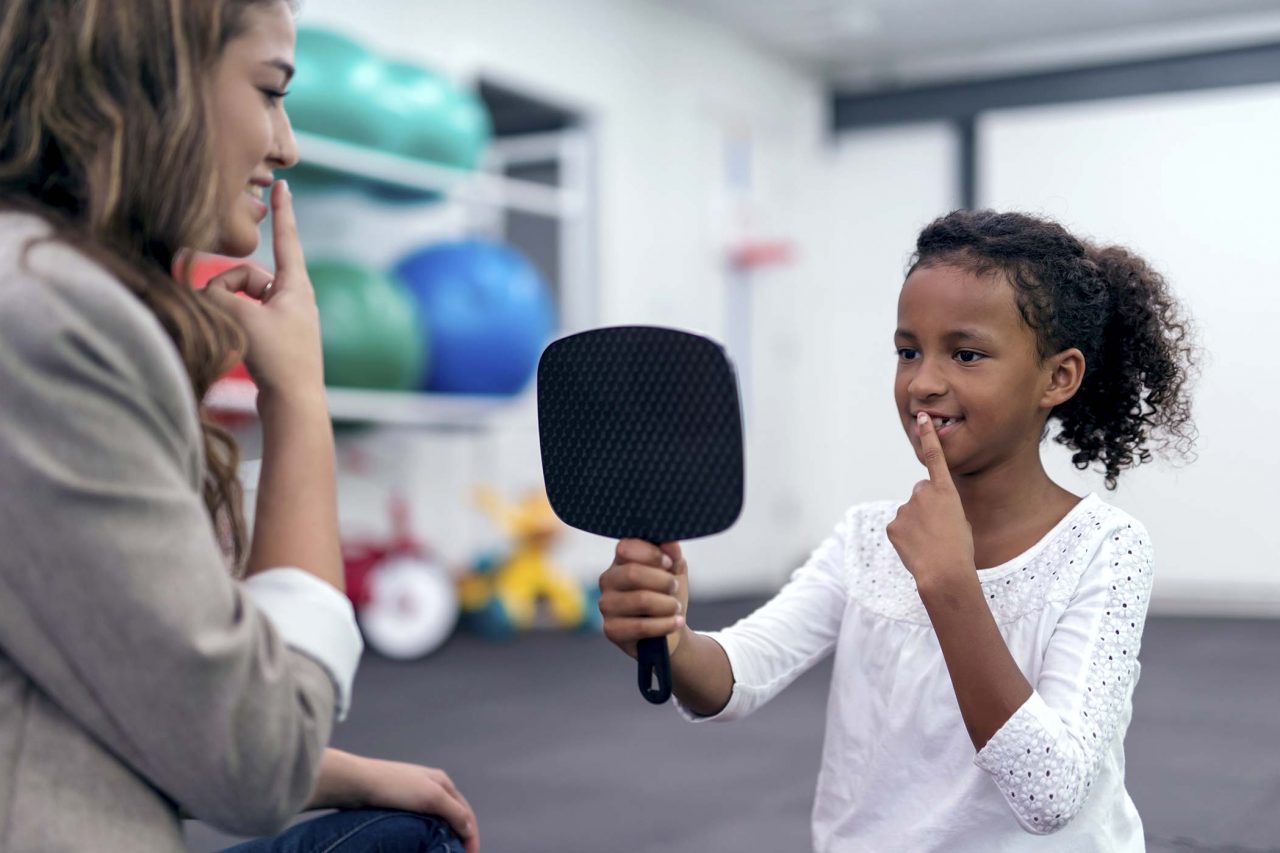 Young girl wearing white shirt holds mirror while practicing speech therapy