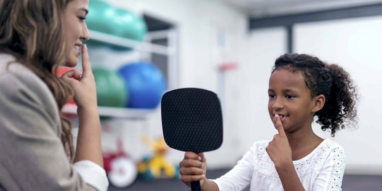 Therapist demonstrates putting index finger to lips as young brown skinned girl in a white top holds up a hand mirror with one hand and copies the gesture with her other hand while looking at her reflection