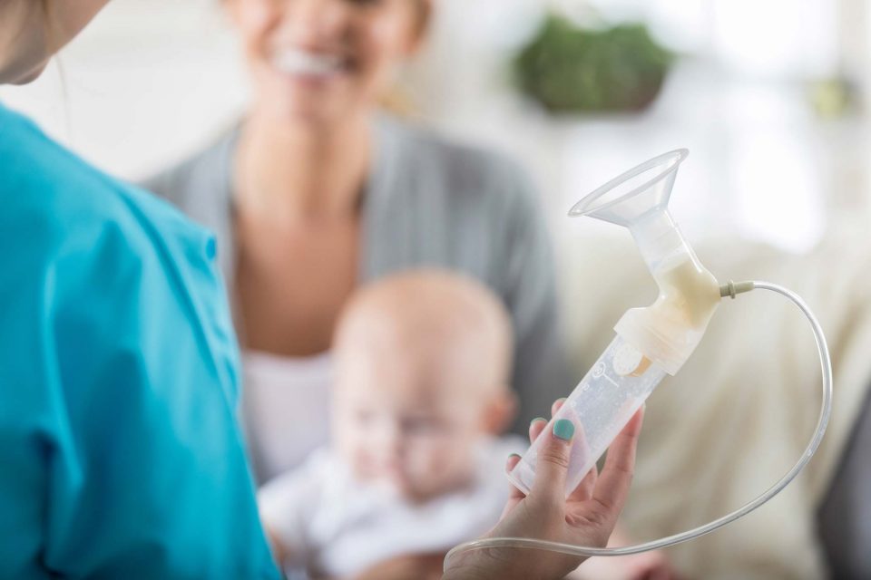 lactation consultant holds up empty plastic breast pump in front of an out of focus client and infant