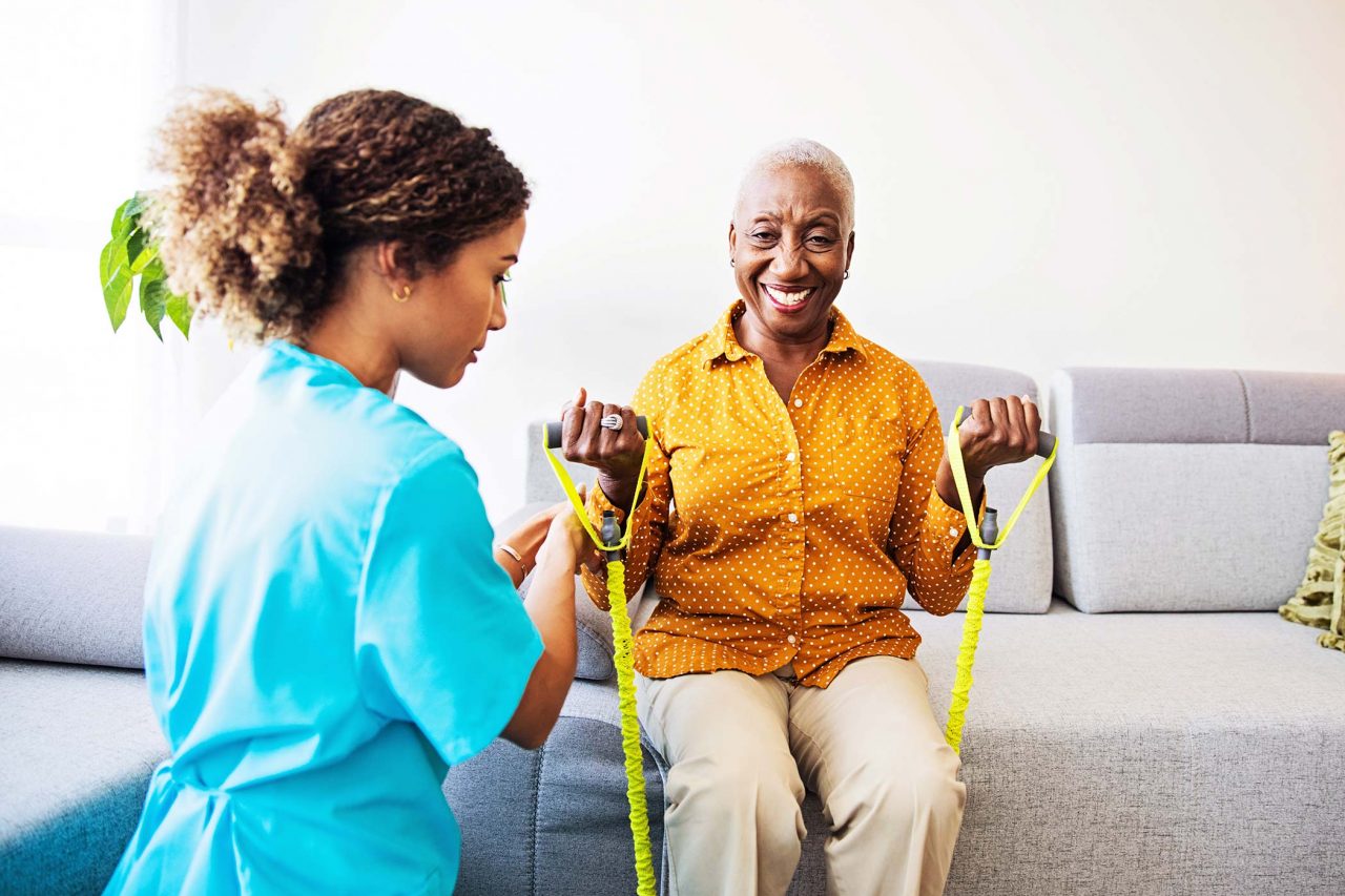 physical therapist in light blue scrubs assists elderly woman with arm strength exercises on couch at home