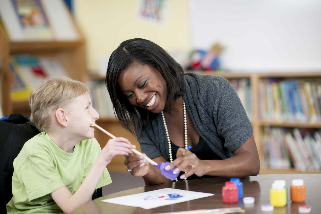 young boy in green shirt dips paintbrush into small jar of purple paint with the help of a smiling therapist