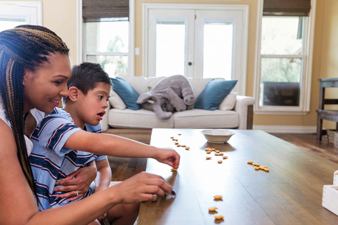 young mom and boy with down syndrome count out goldfish crackers on wooden coffee table