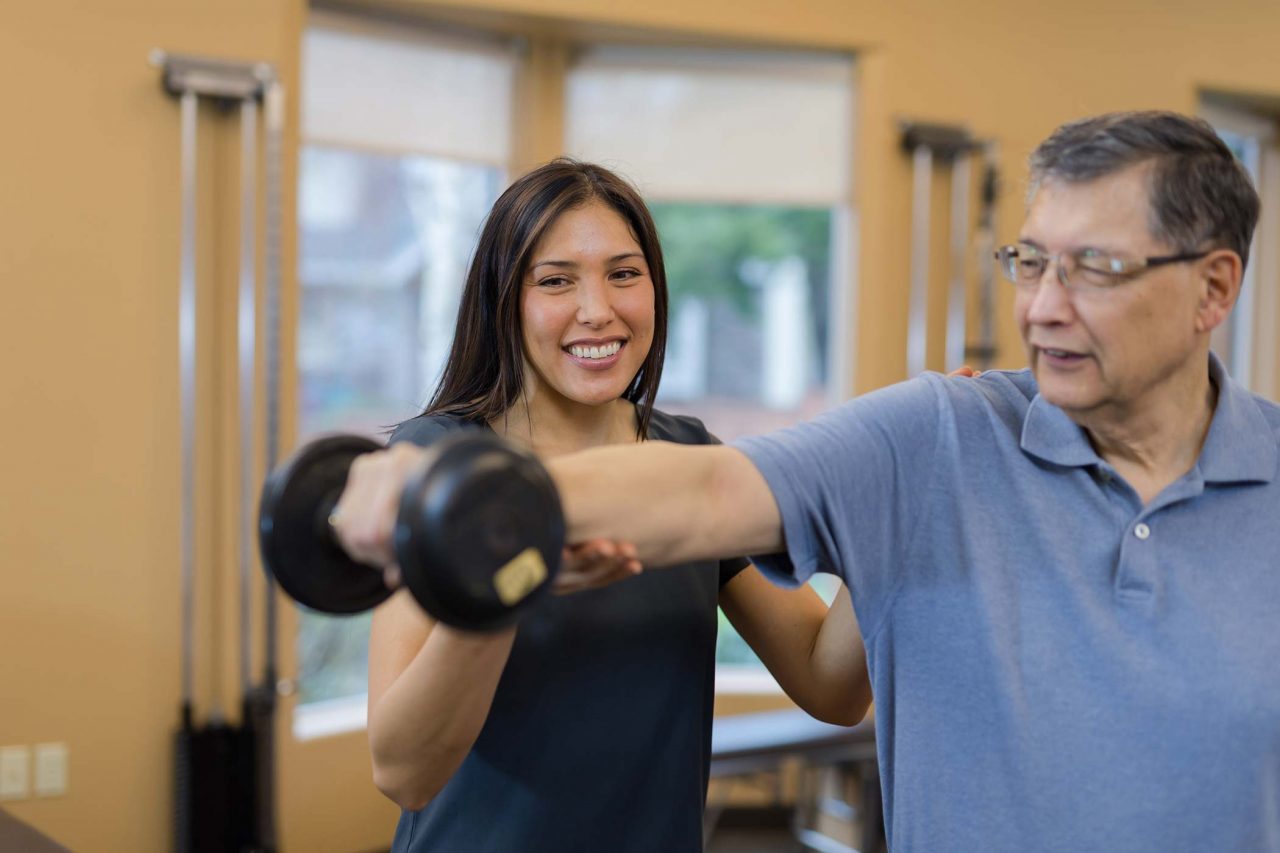 older male patient holds dumbbell straight out from body with help from physical therapist