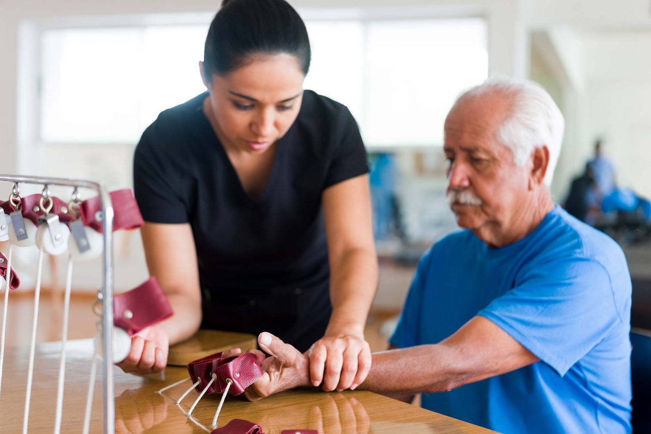 senior male patient sitting and exercising his hand with a physical therapy device next to a occupational therapist