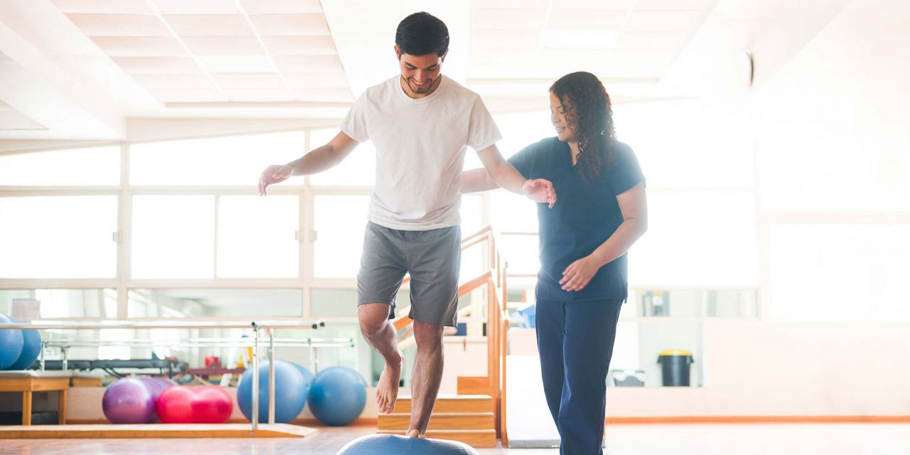 Male in white shirt standing on one leg blue bosu ball getting physical therapy by women in blue scrubs